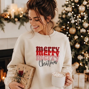 May include: A cream-colored crewneck sweatshirt featuring the text merry merry merry in stacked red and pink script fonts, with the word Christmas written below in black script. The garment is worn while holding a wrapped gift and a mug.