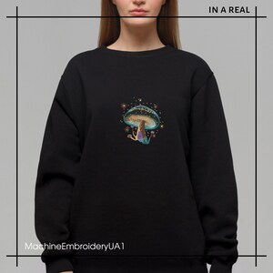May include: Black sweatshirt with a colorful embroidered mushroom design. The mushroom has a blue cap with gold accents and a purple stem. The text "MachineEmbroideryUA1" is at the bottom of the image.