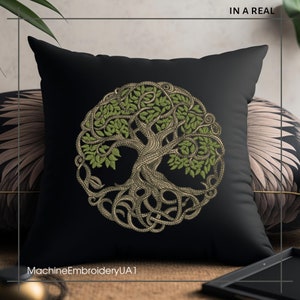 May include: A black pillow with a green and gold embroidered tree of life design. The pillow is on a brown surface with a gold tray and a green plant in the background.