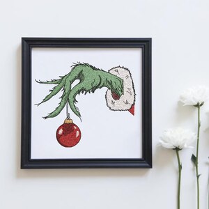 May include: A green Grinch hand holding a red Christmas ornament. The hand is reaching out from a white background.