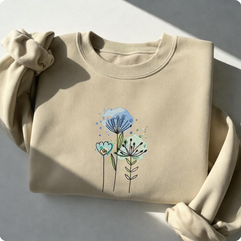 May include: Beige sweatshirt with an embroidered floral design. The design features three stylized flowers in shades of blue and green with delicate stems and leaves. The sweatshirt is folded, with the sleeves visible.