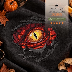 May include: A black fabric featuring a machine embroidery design of a reptilian eye with a bright yellow iris and a vertical black pupil, surrounded by textured red and orange scales, with text reading MACHINE EMBROIDERY DESIGN.