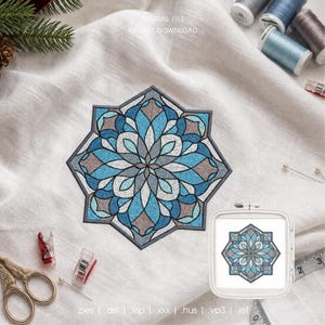 May include: A digital embroidery design featuring a symmetrical, star-shaped mandala pattern in shades of blue, gray, and white. The design is displayed as a stitched sample on fabric and within a square embroidery hoop, accompanied by sewing notions.