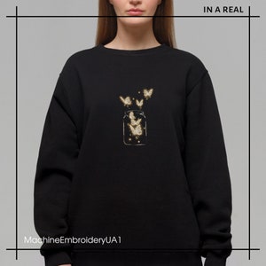 May include: Black sweatshirt with a gold embroidered design of a glass jar with butterflies flying out of it. The text "IN A REAL" is in the top right corner of the image.