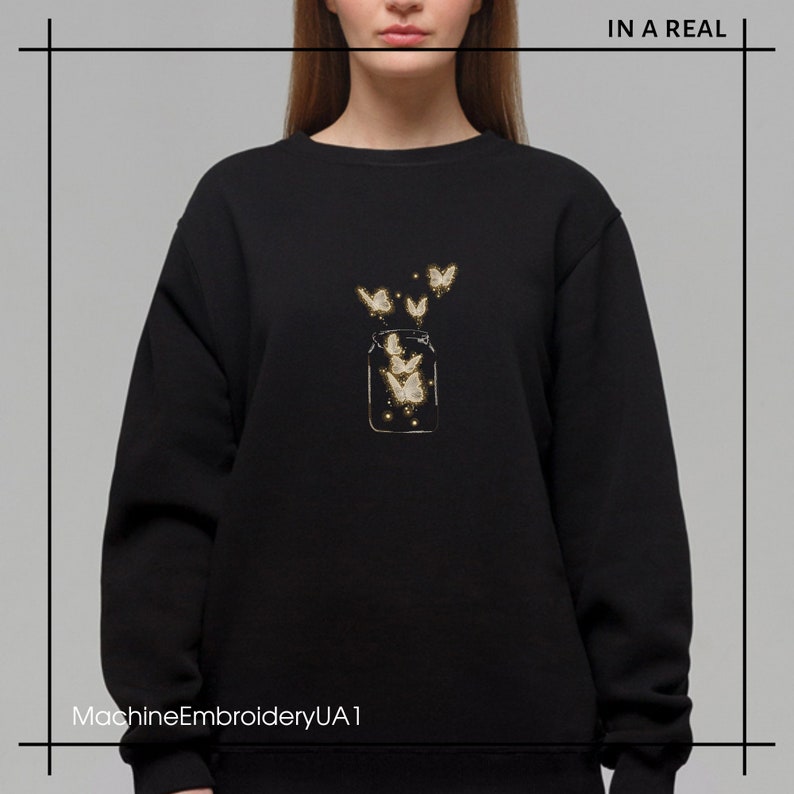 May include: Black sweatshirt with a gold embroidered design of a glass jar with butterflies flying out of it. The text "IN A REAL" is in the top right corner of the image.