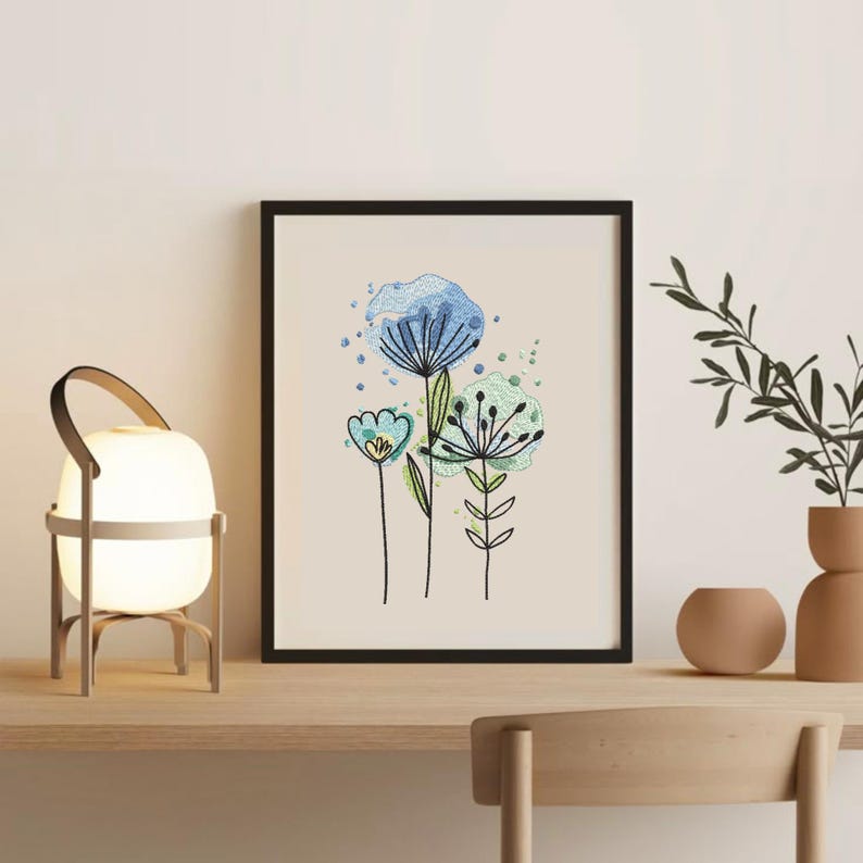 May include: A framed floral art print featuring three stylized flowers in shades of blue and green against a cream background. The artwork is in a black frame and sits on a wooden surface with a lamp and decorative vases.