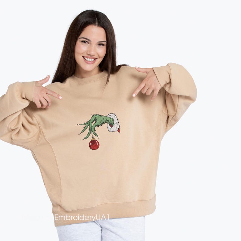 May include: A tan sweatshirt with a green Grinch hand holding a red Christmas ornament. The sweatshirt has the text "EmbroideryUA1" on the bottom.
