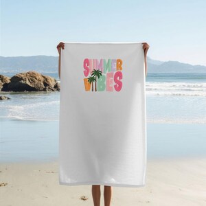 May include: White beach towel with the words "SUMMER VIBES" in colorful, outlined letters. A palm tree graphic is centered below the text. The towel is held up against a beach backdrop with the ocean and mountains visible.