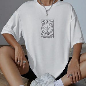 Pu&ograve; includere: Maglietta bianca con una grafica in bianco e nero di una carta dei tarocchi con il segno zodiacale Sagittario.