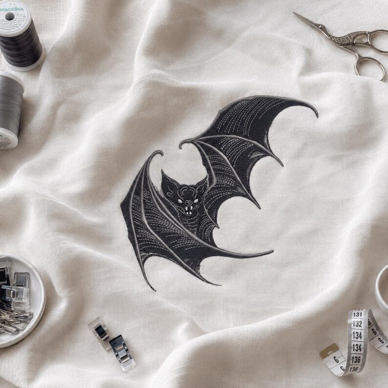 May include: A black embroidered bat design on off-white fabric, surrounded by sewing supplies including two spools of thread, silver scissors, metal clips in a small bowl, and a white measuring tape showing numbers up to 136.