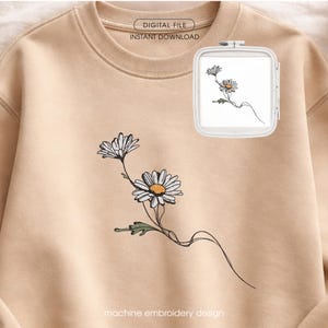 Op de afbeelding: Beige sweatshirt met een madeliefjes borduurontwerp. Het ontwerp toont twee witte madeliefjes met gele centra en groene bladeren. De woorden "DIGITAL FILE INSTANT DOWNLOAD" en "machine embroidery design" zijn gedrukt.