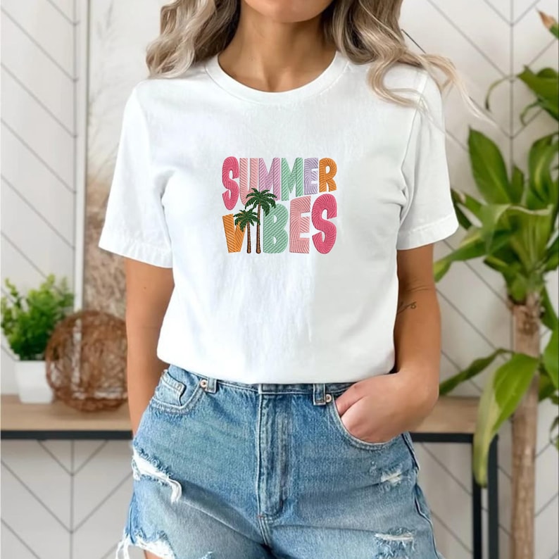 May include: White t-shirt with the text "SUMMER VIBES" in colorful, embroidered letters. The design includes a palm tree graphic. The shirt is paired with denim shorts.
