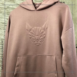 K&ouml;nnte beinhalten: Ein staubiger rosa Hoodie mit einem geometrischen Katzendesign, das auf der Vorderseite gestickt ist. Der Text "IN A REAL" ist in der oberen rechten Ecke des Bildes sichtbar.