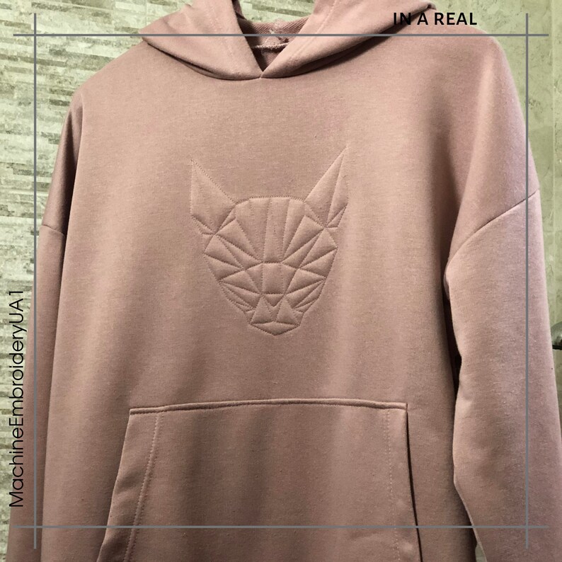 K&ouml;nnte beinhalten: Ein staubiger rosa Hoodie mit einem geometrischen Katzendesign, das auf der Vorderseite gestickt ist. Der Text "IN A REAL" ist in der oberen rechten Ecke des Bildes sichtbar.