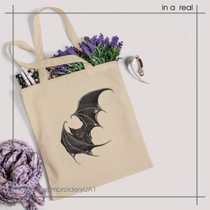 May include: A beige tote bag featuring a black graphic illustration of a bat with spread wings. The bag contains purple lavender sprigs and white pens. The text "in a real" appears in the top right corner, and "MachineEmbroideryUA1" is at the bottom.