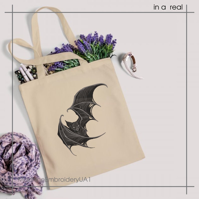May include: A beige tote bag featuring a black graphic illustration of a bat with spread wings. The bag contains purple lavender sprigs and white pens. The text "in a real" appears in the top right corner, and "MachineEmbroideryUA1" is at the bottom.