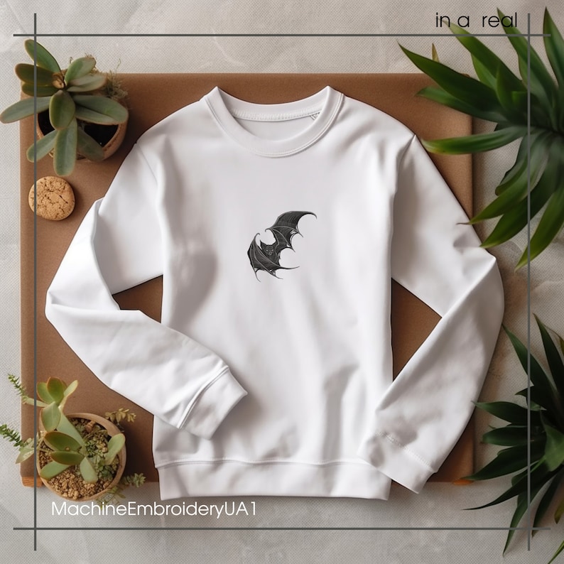 May include: A white long-sleeve sweatshirt featuring a black embroidered bat graphic centered on the chest. The garment has a crew neckline, ribbed cuffs, and a ribbed hem. The text "in a real" and "MachineEmbroideryUA1" appear in the corners.