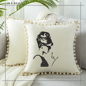 May include: A white throw pillow with a black embroidered image of a woman with a bun hairstyle and sunglasses. The pillow has a white pom-pom trim.