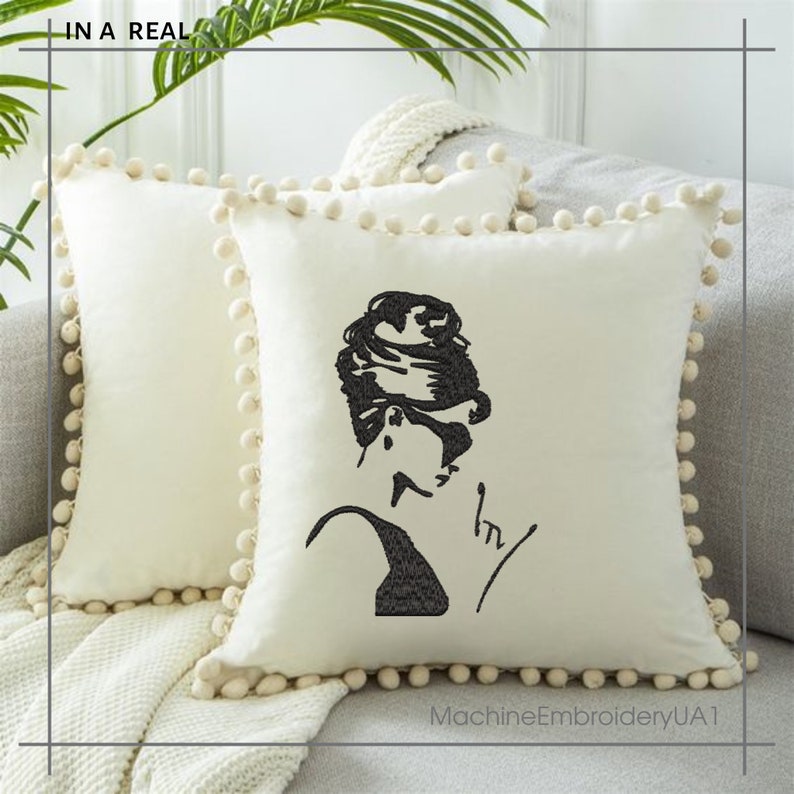 May include: A white throw pillow with a black embroidered image of a woman with a bun hairstyle and sunglasses. The pillow has a white pom-pom trim.
