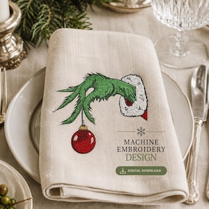Könnte beinhalten: Eine beige Leinen-Serviette mit einer Stickerei einer grünen, pelzigen Hand mit weißem Weihnachtsmann-Ärmelaufschlag, die eine rote Weihnachtskugel hält. Der Text lautet MACHINE EMBROIDERY DESIGN und DIGITAL DOWNLOAD mit Schneeflocke.