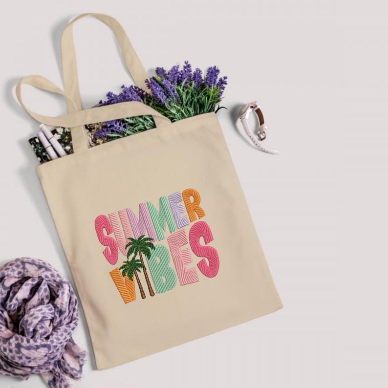 May include: Beige tote bag with the embroidered text "SUMMER VIBES" in colorful, block letters, and two palm trees. The bag is filled with lavender and other items. A scarf and a watch are also visible.