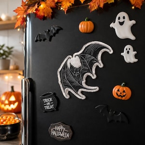 May include: A black and white embroidered bat patch with detailed wing patterns, displayed on a black refrigerator alongside various Halloween-themed magnets including pumpkins, ghosts, bats, and signs reading Trick or Treat and Happy Halloween.