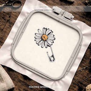 Può includere: Design di ricamo a macchina con una margherita con petali bianchi e centro giallo, attaccata a una spilla da balia. Il design è incorniciato in un telaio da ricamo grigio, con le parole "DIGITAL FILE MACHINE EMBROIDERY" in alto.