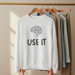 Może przedstawiać: Biała bluza z okrągłym dekoltem z czarną grafiką m&oacute;zgu i napisem "USE IT". Bluza wisi na wieszaku z innymi ubraniami. Projekt jest prosty i odważny.