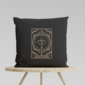 Pu&ograve; includere: Cuscino nero con un designo dorato del segno zodiacale Sagittario. Il design presenta una freccia e un arco all'interno di un cerchio con stelle e lune. Il testo "SAGITTARIO" &egrave; sotto il design.