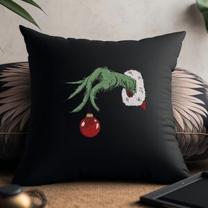 May include: Black throw pillow with an embroidered green hand holding a red Christmas ornament. The hand is wearing a white fur cuff.
