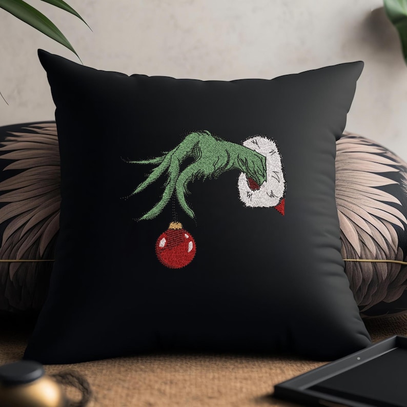 May include: Black throw pillow with an embroidered green hand holding a red Christmas ornament. The hand is wearing a white fur cuff.