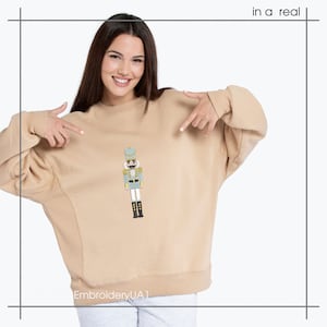 May include: A tan sweatshirt with a detailed embroidered image of a nutcracker soldier on the front. The nutcracker is in shades of blue, gold, and black. The text "in a real" is in the upper right corner. The Etsy shop name "EmbroideryUA1" is at the bottom.