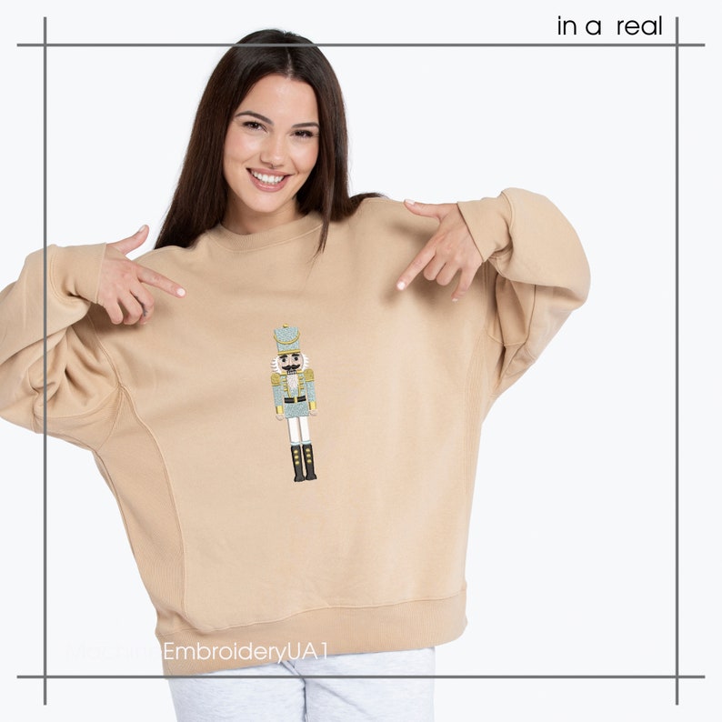 May include: A tan sweatshirt with a detailed embroidered image of a nutcracker soldier on the front. The nutcracker is in shades of blue, gold, and black. The text "in a real" is in the upper right corner. The Etsy shop name "EmbroideryUA1" is at the bottom.