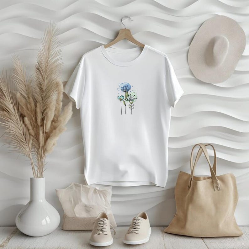 May include: White t-shirt with a floral design in blue and green. The shirt is hanging on a wooden hanger. Beige sneakers, a tote bag, and a hat are also in the image, suggesting a casual, everyday style.