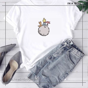 May include: White t-shirt with a graphic of The Little Prince sitting on an asteroid with a fox. The text "IN A REAL" is in the top right corner of the image.