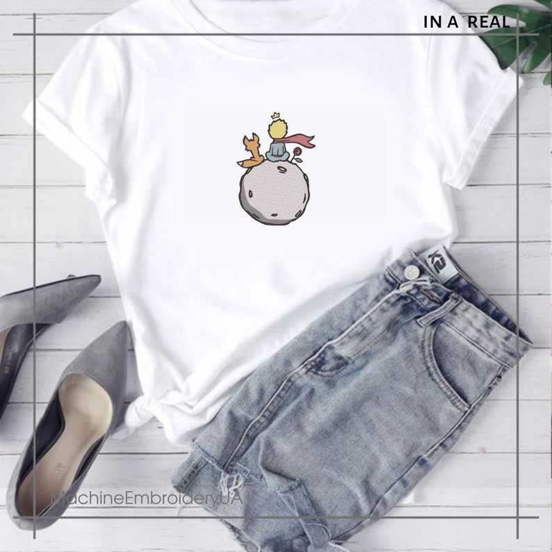 May include: White t-shirt with a graphic of The Little Prince sitting on an asteroid with a fox. The text "IN A REAL" is in the top right corner of the image.