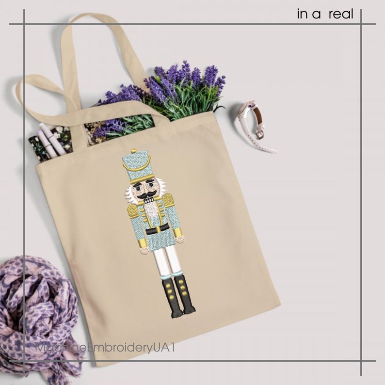 May include: Beige tote bag featuring an embroidered nutcracker design in light blue, gold, and black. The bag contains lavender and pens. The text "in a real" is in the upper right corner. The text "MachineEmbroideryUAT" is at the bottom.