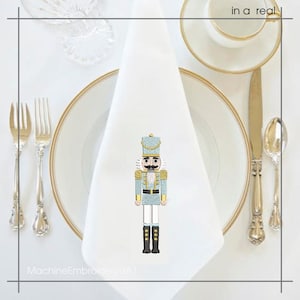 May include: A white cloth napkin with a light blue and gold embroidered nutcracker design. The napkin is placed on a white plate with a gold rim, alongside silverware and a teacup.