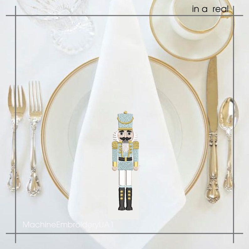 May include: A white cloth napkin with a light blue and gold embroidered nutcracker design. The napkin is placed on a white plate with a gold rim, alongside silverware and a teacup.