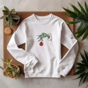 May include: White sweatshirt with a green Grinch hand holding a red Christmas ornament. The hand is reaching out from the sweatshirt.