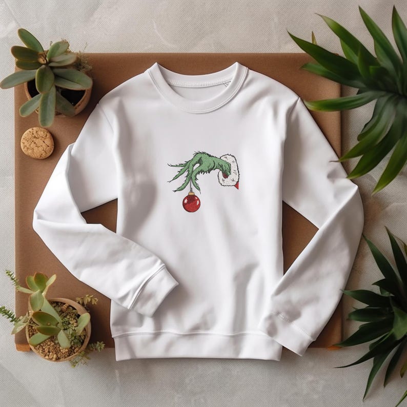 May include: White sweatshirt with a green Grinch hand holding a red Christmas ornament. The hand is reaching out from the sweatshirt.
