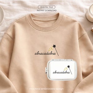 Può includere: Una felpa beige con la parola "abracadabra" in corsivo, accompagnata da un disegno di bacchetta magica e stella. È visibile anche un telaio da ricamo bianco con lo stesso disegno. Sono presenti anche le parole "DIGITAL FILE INSTANT DOWNLOAD" e "machine embroidery design".