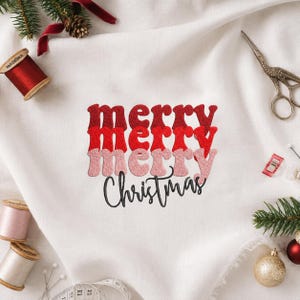 May include: White fabric featuring an embroidered design with the word merry repeated three times in stacked, textured red and pink letters, followed by the word Christmas in black cursive script, surrounded by sewing supplies.