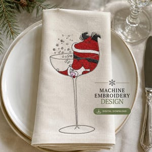 Könnte beinhalten: Eine cremefarbene Stoffserviette mit einer Stickerei, die den Weihnachtsmann kopfüber in einem großen Cocktailglas mit Stiel zeigt, umgeben von kleinen gestickten Schneeflocken und Blasen, mit dem Text MACHINE EMBROIDERY DESIGN.