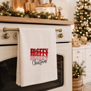 May include: A cream-colored kitchen towel hanging on a white oven handle. The towel features the text 'merry' in stacked red and pink script, followed by the word 'Christmas' in black cursive lettering centered on the fabric.
