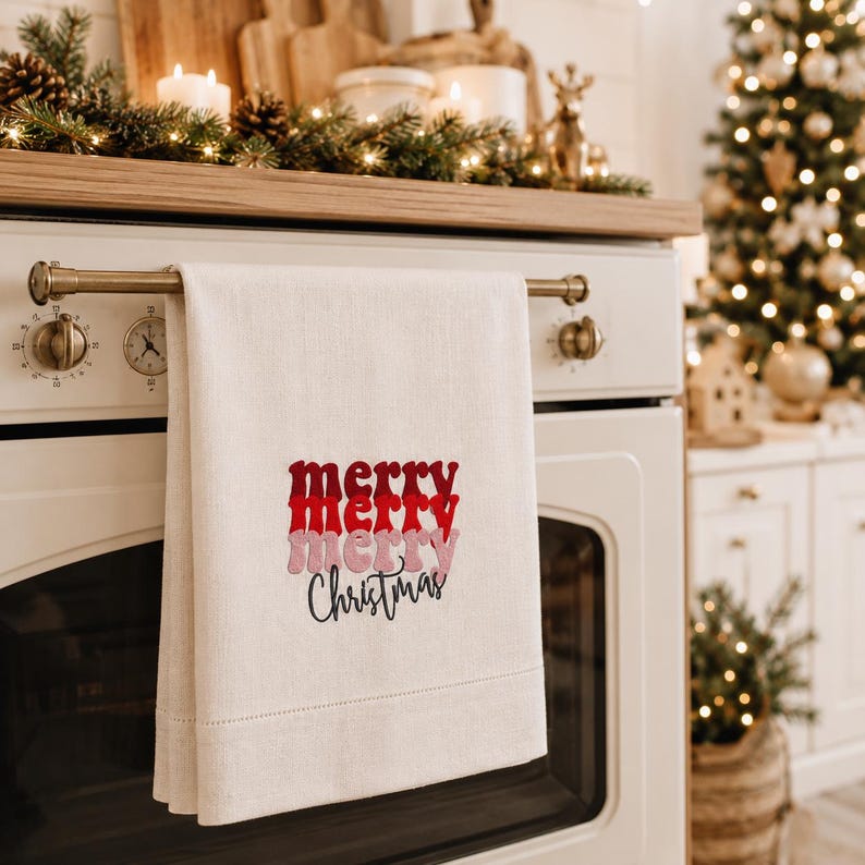 May include: A cream-colored kitchen towel hanging on a white oven handle. The towel features the text 'merry' in stacked red and pink script, followed by the word 'Christmas' in black cursive lettering centered on the fabric.