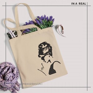 May include: A beige tote bag with a black silhouette of a woman with her hair in a bun. The woman is wearing a black top and has a signature below her. The text "In a Real" is in the top right corner of the image.