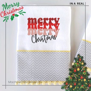 May include: A white kitchen towel featuring embroidered text that reads merry merry merry Christmas in stacked red and pink script. The towel has a gray and white chevron patterned border at the bottom with a yellow trim and small yellow pom-poms.