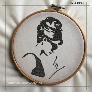 May include: Black and white embroidered image of a woman's face in a circular wooden embroidery hoop. The text "IN A REAL" is at the top of the hoop and the text "MachineEmbroideryUA1" is at the bottom.
