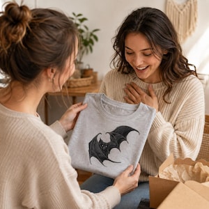 May include: A light gray crew neck t-shirt featuring a detailed black graphic illustration of a bat with spread wings centered on the front. The bat design includes intricate line work and shading, showcasing the creature in a flying position.